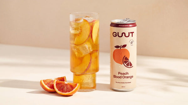 GUUT Peach Blood Orange prebiotic soda styled with fresh orange fruit ingredients on a clean background.